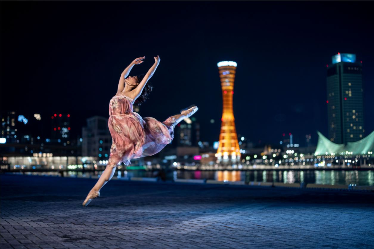 Ballerina leaping, arms outstretched, with city lights in the background, photographed using the Canon Speedlite EL-1 Flash