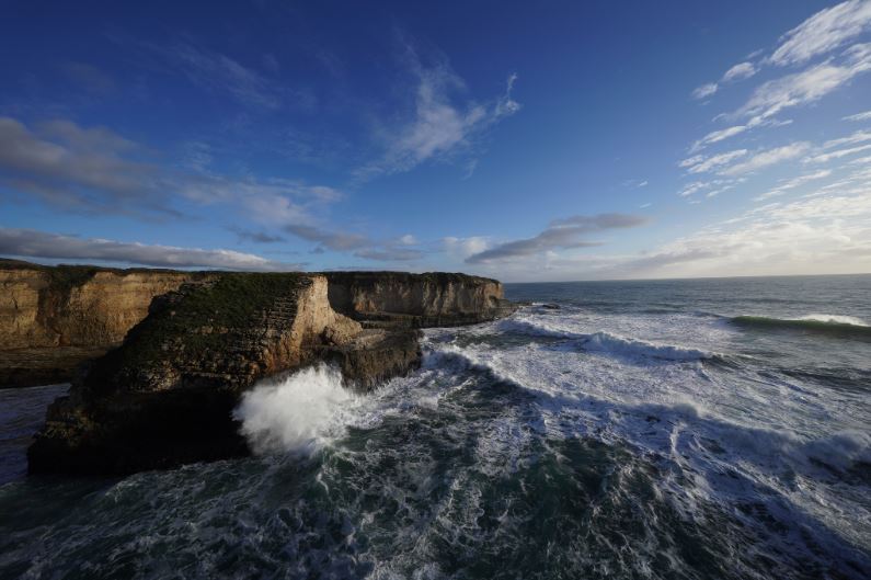 Ocean waves breaking on rocky cliffs beneath a cloudy blue sky, photographed with the Sony 12-24mm f2.8 GM lens