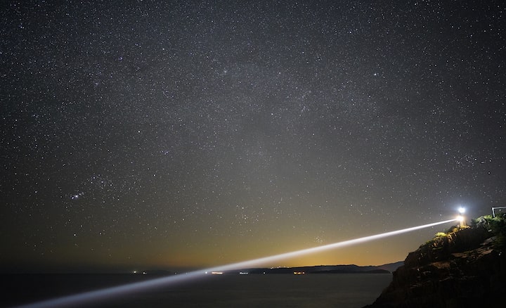 Light from a lighthouse beaming over the sea below a starry night sky, photographed with the Sony 24mm 1.4 GM lens