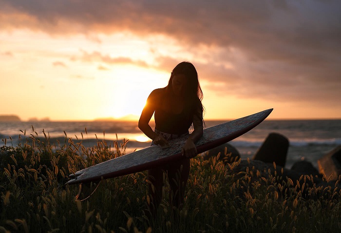 Woman waxing a surfboard among tall grass, sun setting over the ocean behind, shot with the Canon RF 50mm f1.2L USM lens