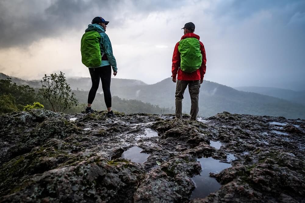 A woman and a man, both in windbreakers are standing beside a mountain cliff