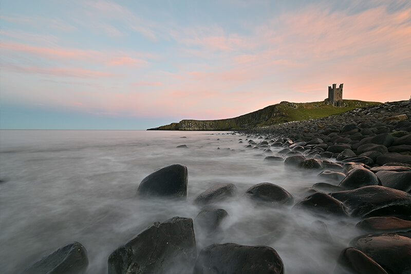 Ocean waves washing over rocks, with a castle atop a green hill in the distance, photographed with the Nikon Z 14-30mm lens