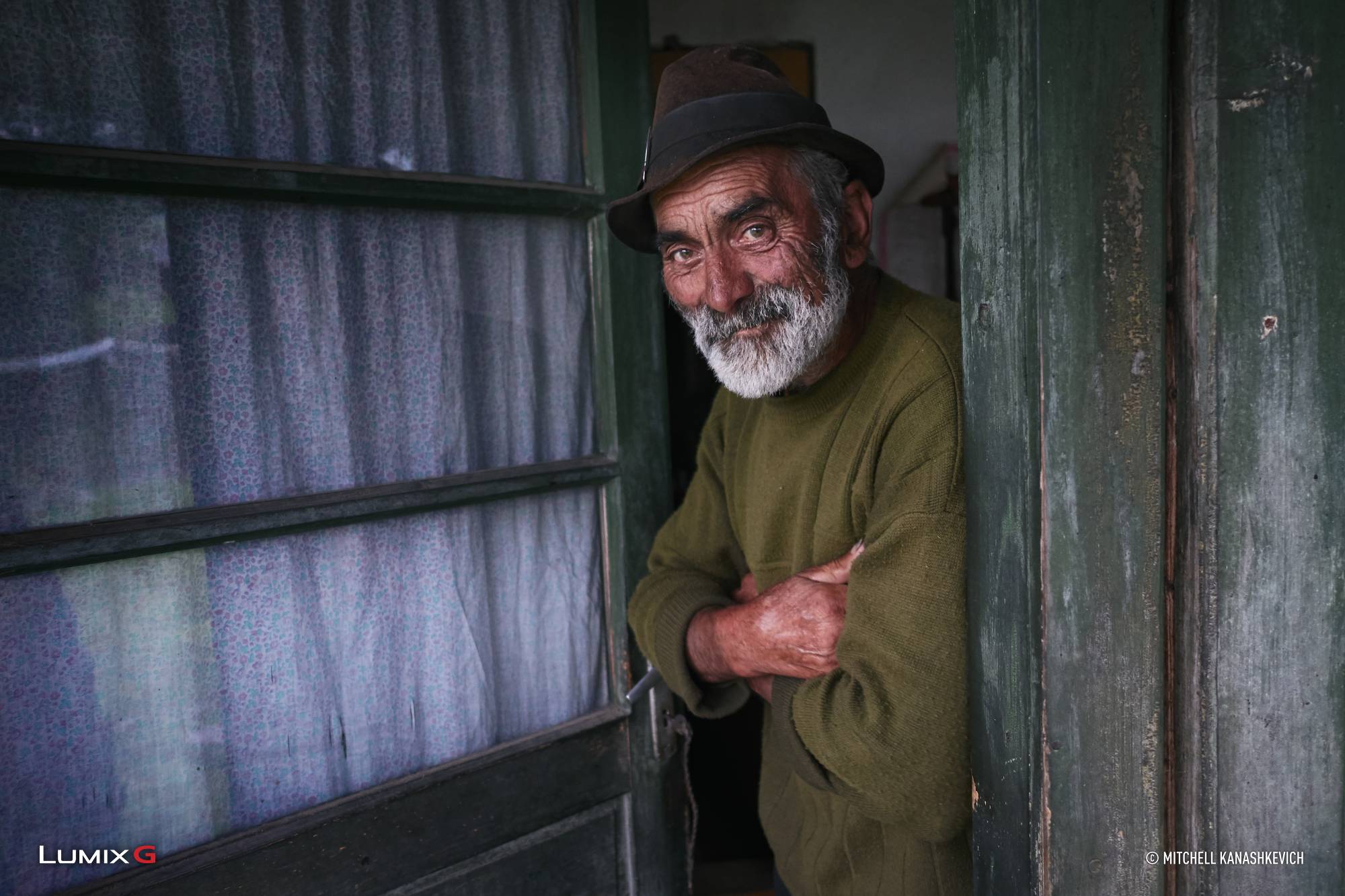 Grey-bearded man in a brown hat smiling in a rustic green doorway, photographed with the Panasonic LUMIX G85 camera