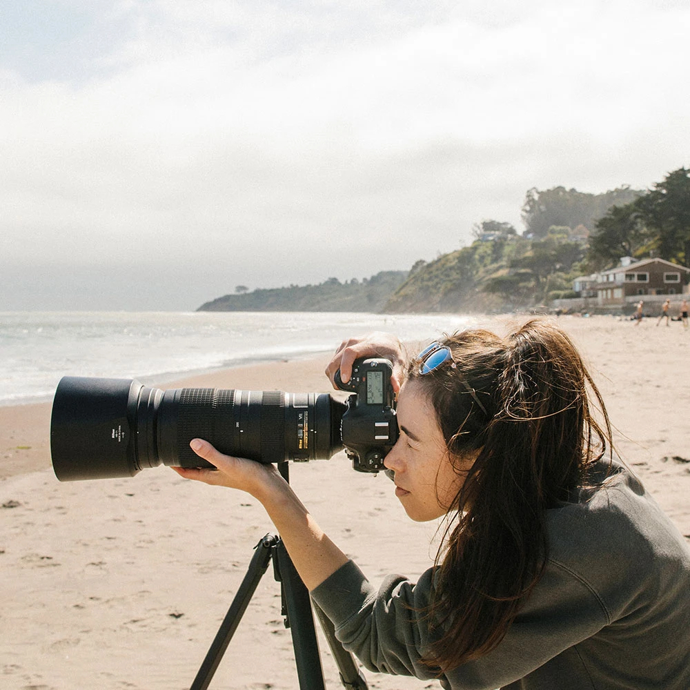 Woman photographing on the beach using a Nikon DSLR camera and telephoto lens on the Peak Design Travel Tripod