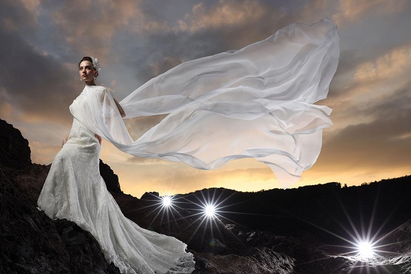 Bride in white lace gown, veil blowing in the wind, standing on rocks below a cloudy sunset, shot using Canon RF 28-70mm f2L