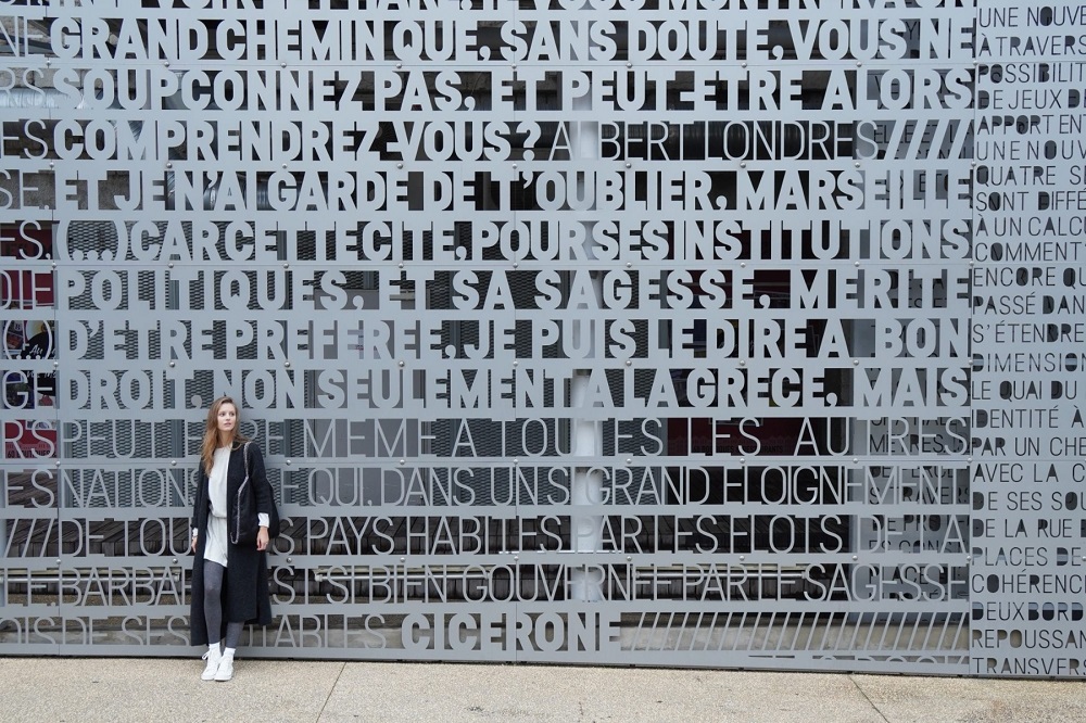 Young woman in long dark coat posing in front of a wall displaying French words, photographed with the Sony a6400