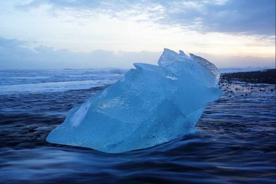 Lump of blue ice sitting in ocean shallows with a cloudy sky in the background, shot with the Sony FE 24-70mm f/2.8 GM Lens