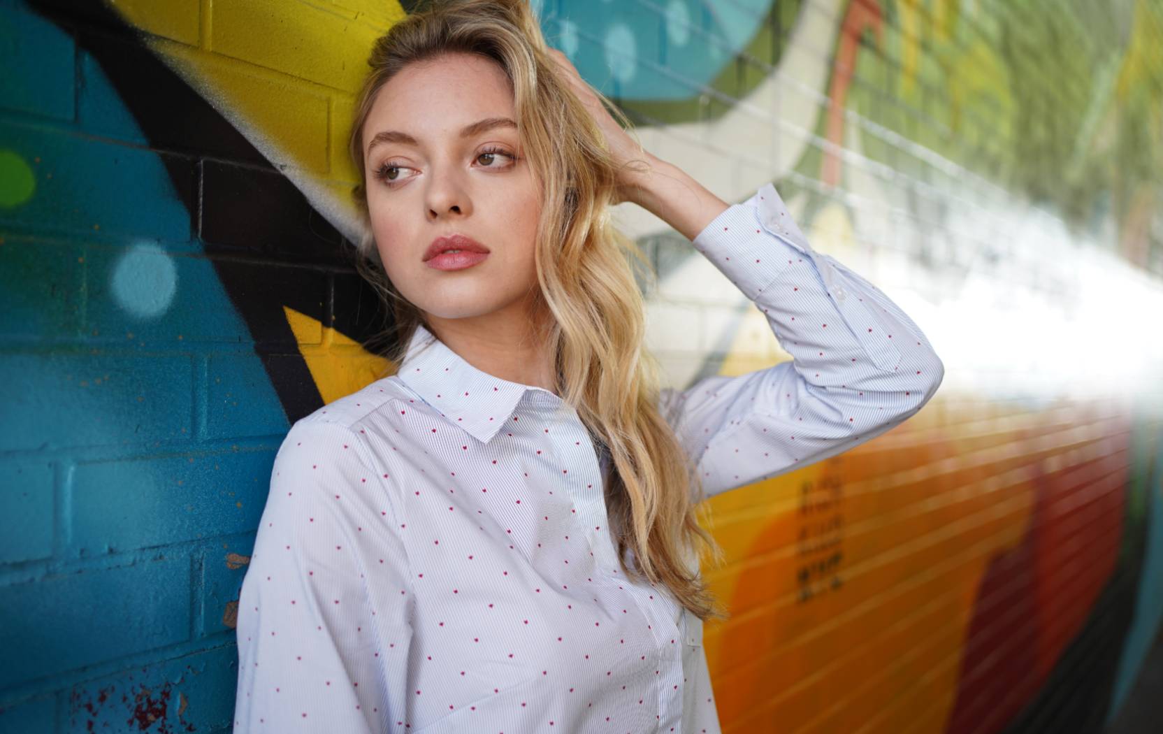 Young blonde woman in collared shirt resting against a colourful graffiti mural, photographed with the Sony FE 35mm f1.8 lens