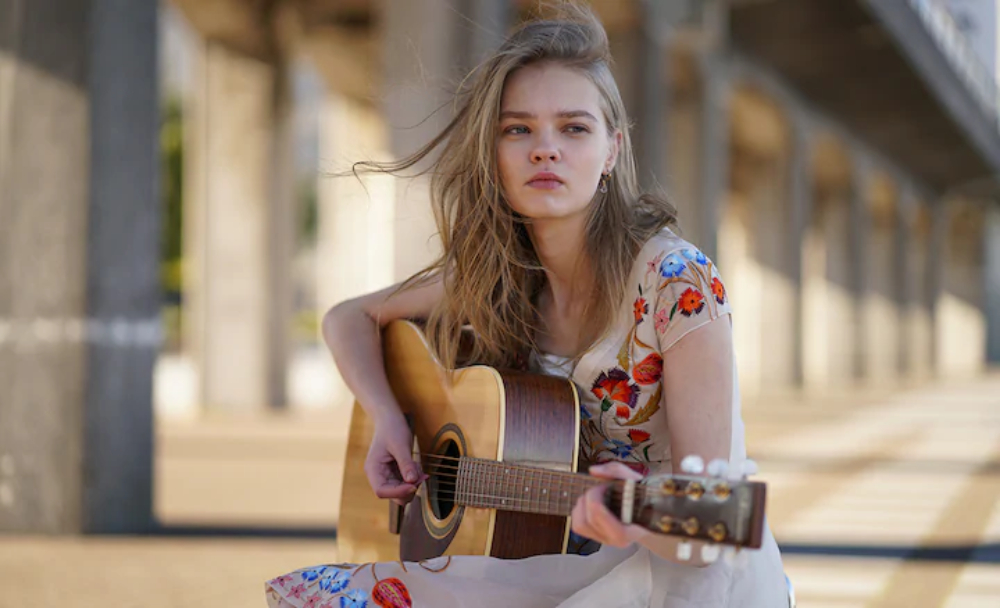 Young blonde-haired woman playing acoustic guitar with concrete bridge in the background, shot with the Sony 85mm 1.8 lens