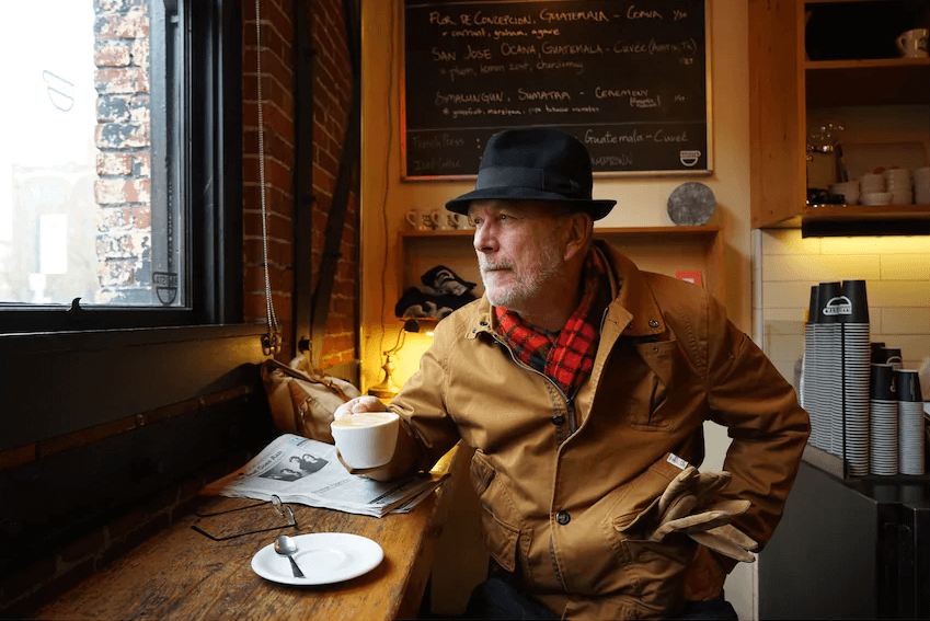 Man in jacket and hat holding a cup of coffee while seated inside a cafe, photographed with the Sony 24 to 70mm f4 Zeiss lens