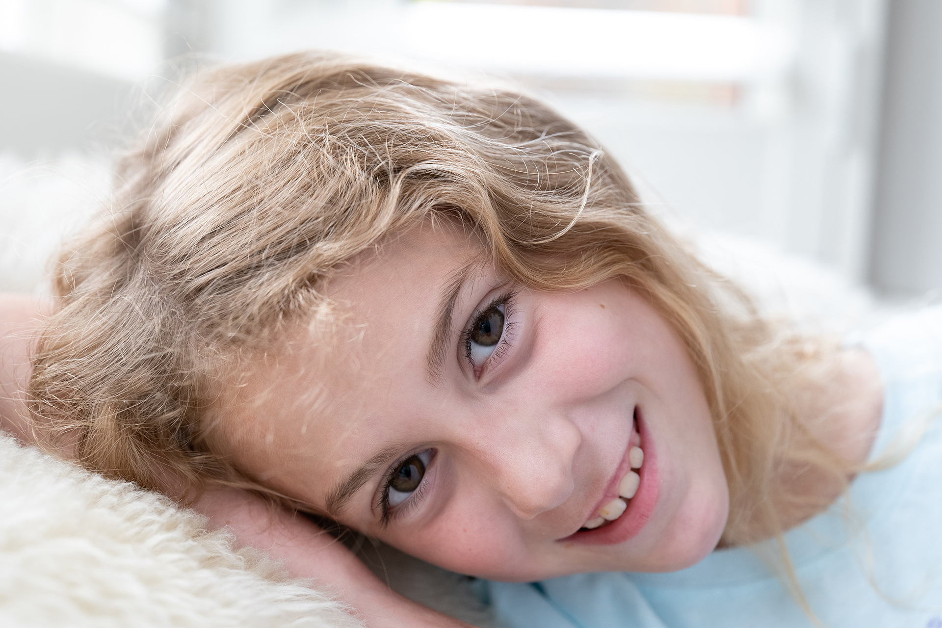 High key portrait of girl with blonde hair resting against shaggy white blanket, photographed using Panasonic Lumix S PRO 24-70mm f2.8 lens