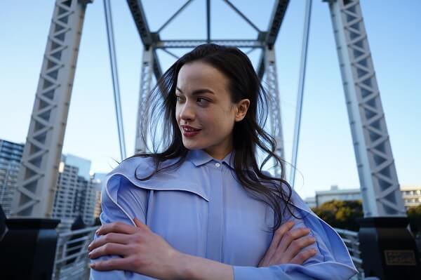 Young smiling woman with arms folded, standing before a tall metal structure, photographed with the Sony 24mm f2.8 G lens